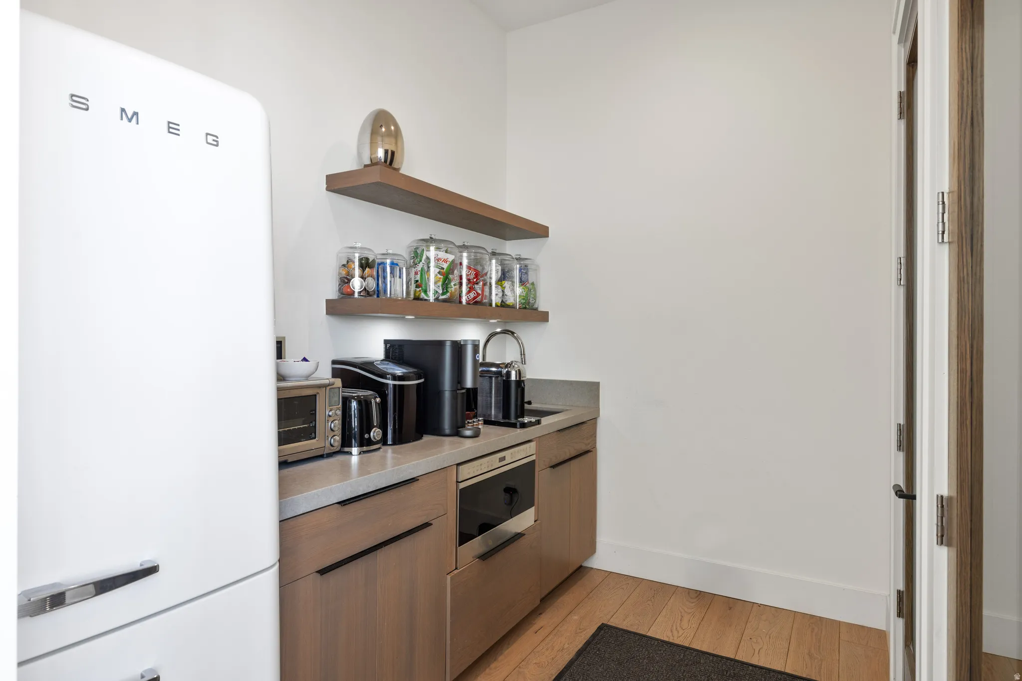 Integrated beverage station featuring a compact refrigerator, wood-finish cabinetry, light-toned countertop, and open wood shelving