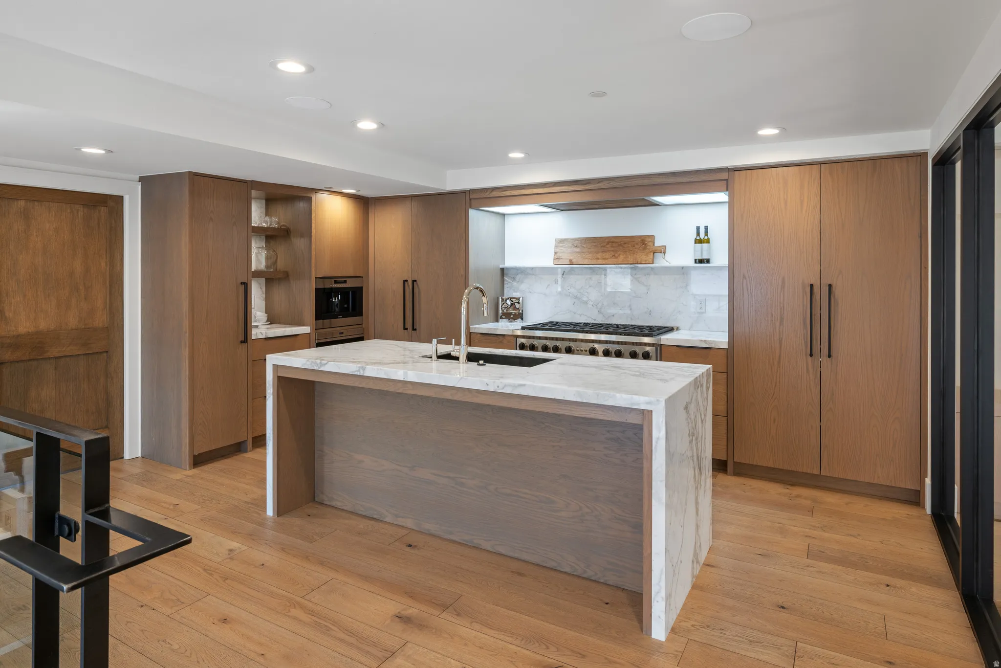 Gourmet kitchen featuring a large central island with waterfall-edge countertop, premium stainless steel range, full-height marble backsplash, custom wood cabinetry, and recessed lighting