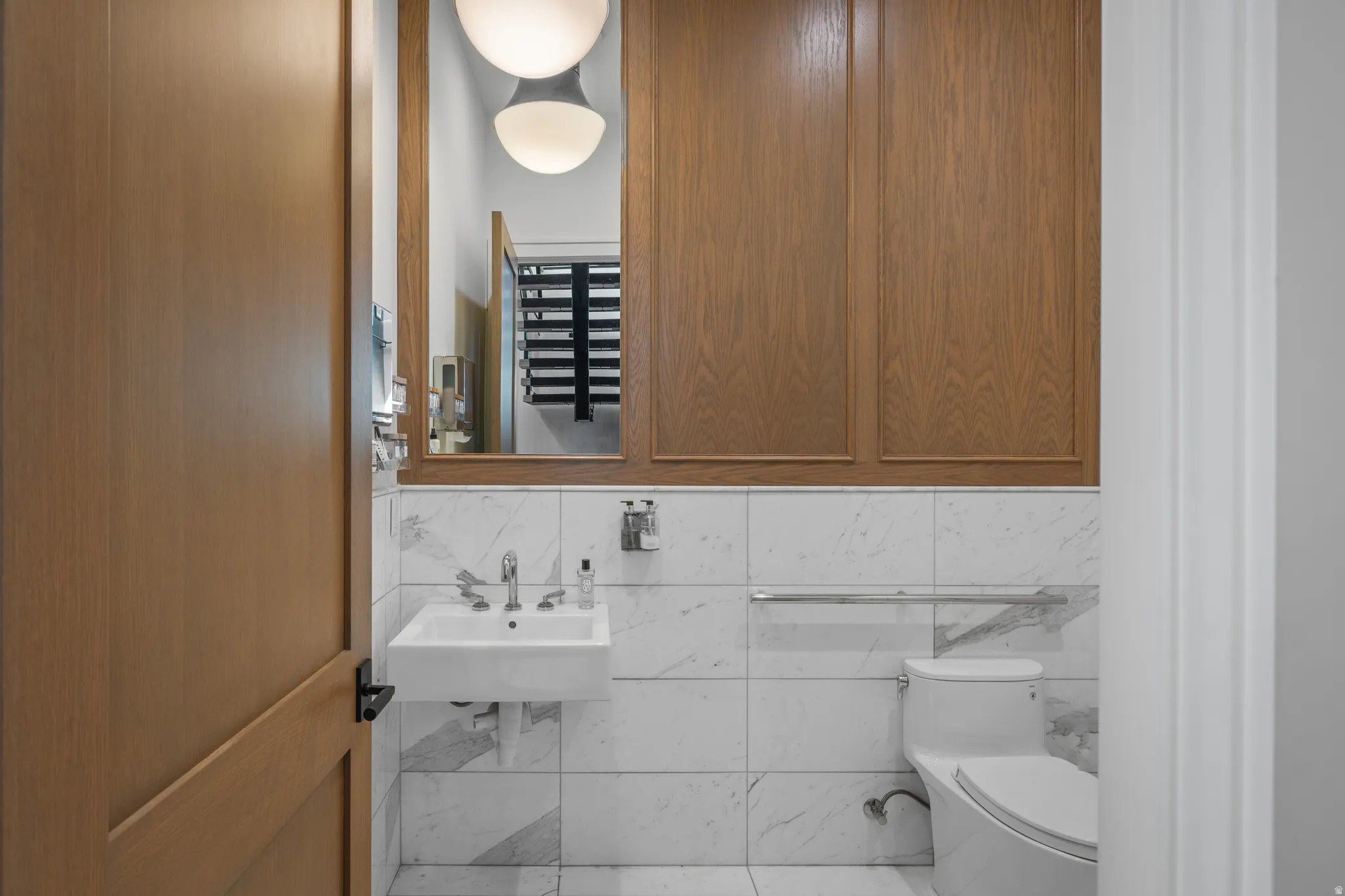 Powder room featuring a wall-mounted sink, a toilet, and wood-paneled walls above white marble-look wall tiles