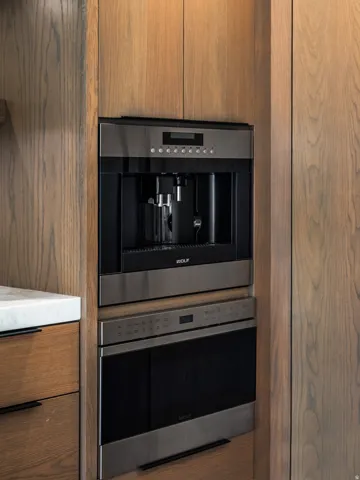 Integrated stainless steel built-in coffee system with a complementing wall oven, set within custom wood cabinetry with black linear hardware and a white stone countertop