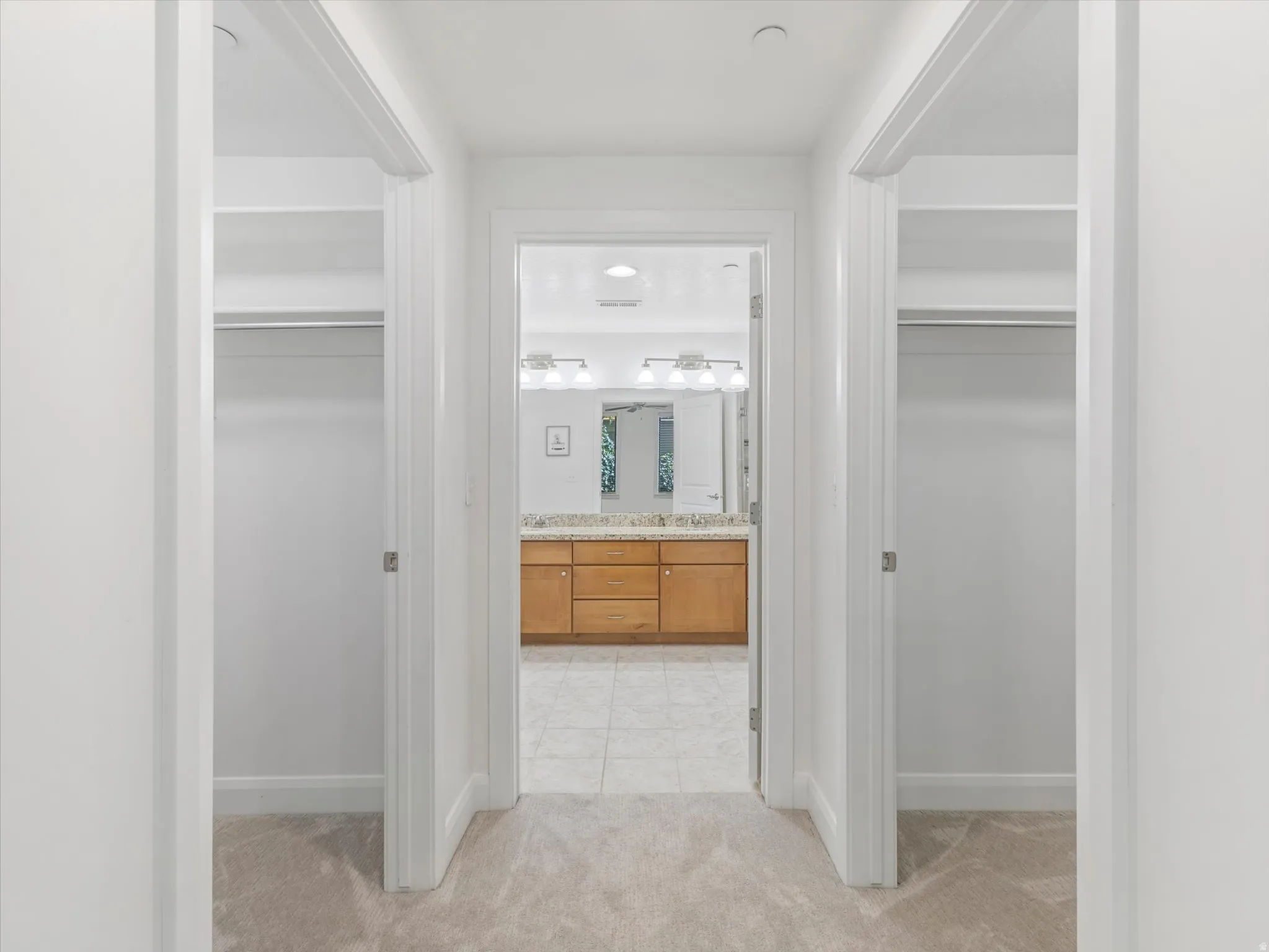Hallway with light-toned carpet flooring, leading to a bathroom featuring a dual vanity with a light wood-finish cabinet and a light-toned countertop