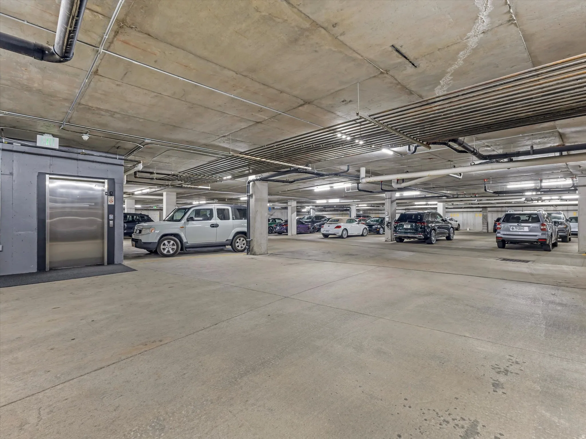 Underground parking garage featuring concrete flooring, concrete support columns, overhead lighting, exposed ceiling pipes, and a stainless steel elevator