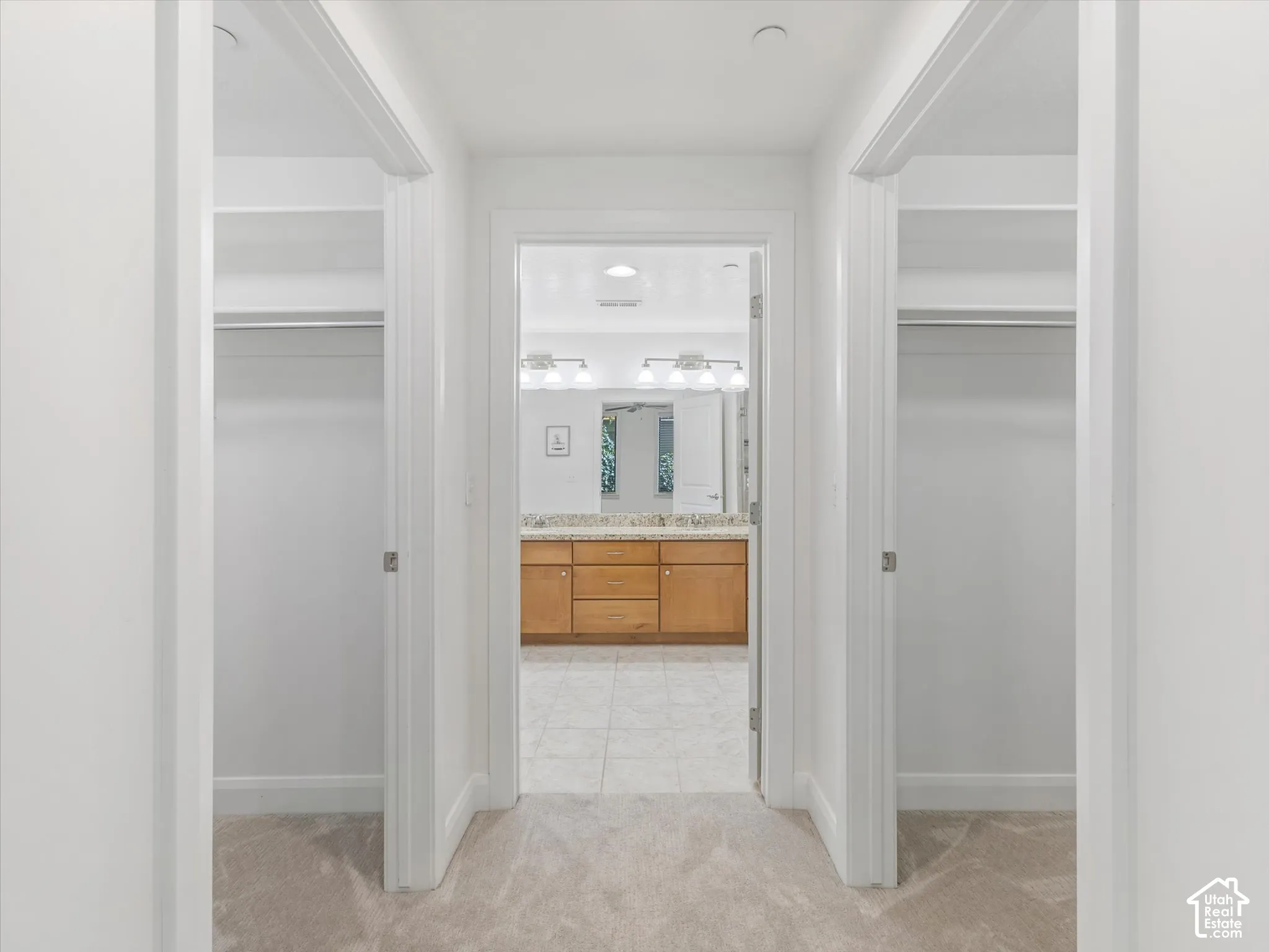 Hallway with light-toned carpet flooring, leading to a bathroom featuring a dual vanity with a light wood-finish cabinet and a light-toned countertop