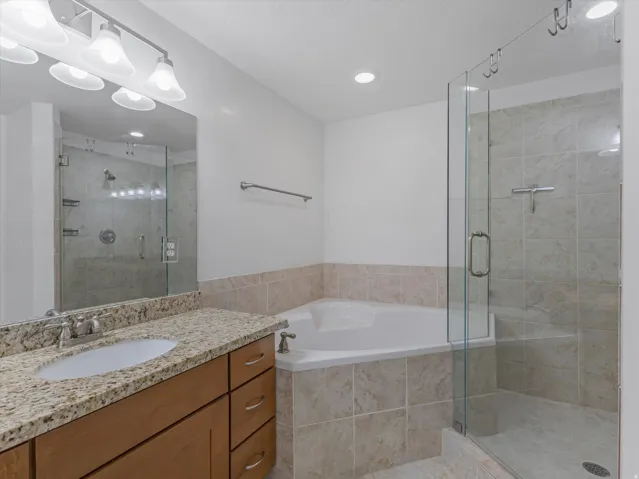 Bathroom featuring a corner soaking tub, glass-enclosed shower with tiled walls, and a vanity with a granite-style countertop