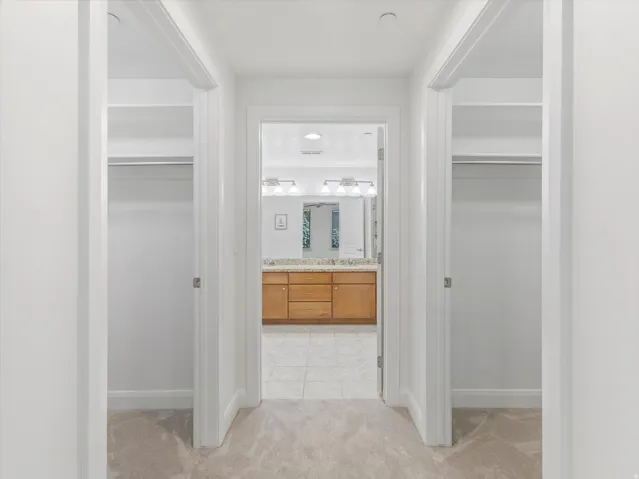 Hallway with light-toned carpet flooring, leading to a bathroom featuring a dual vanity with a light wood-finish cabinet and a light-toned countertop