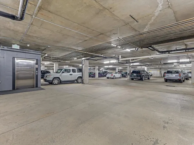 Underground parking garage featuring concrete flooring, concrete support columns, overhead lighting, exposed ceiling pipes, and a stainless steel elevator