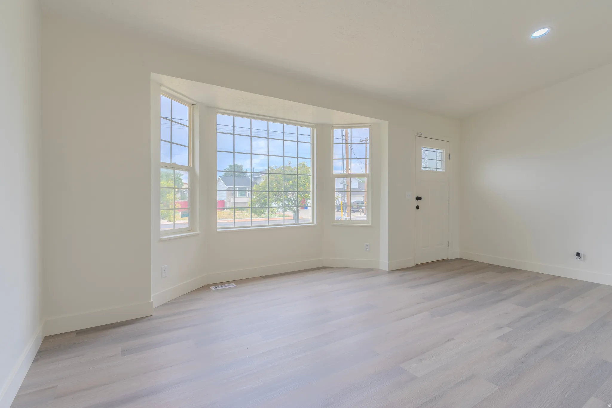 Spacious room featuring light wood-finish flooring, a prominent bay window with grid patterns, recessed ceiling lighting, and a white entry door with glass panels
