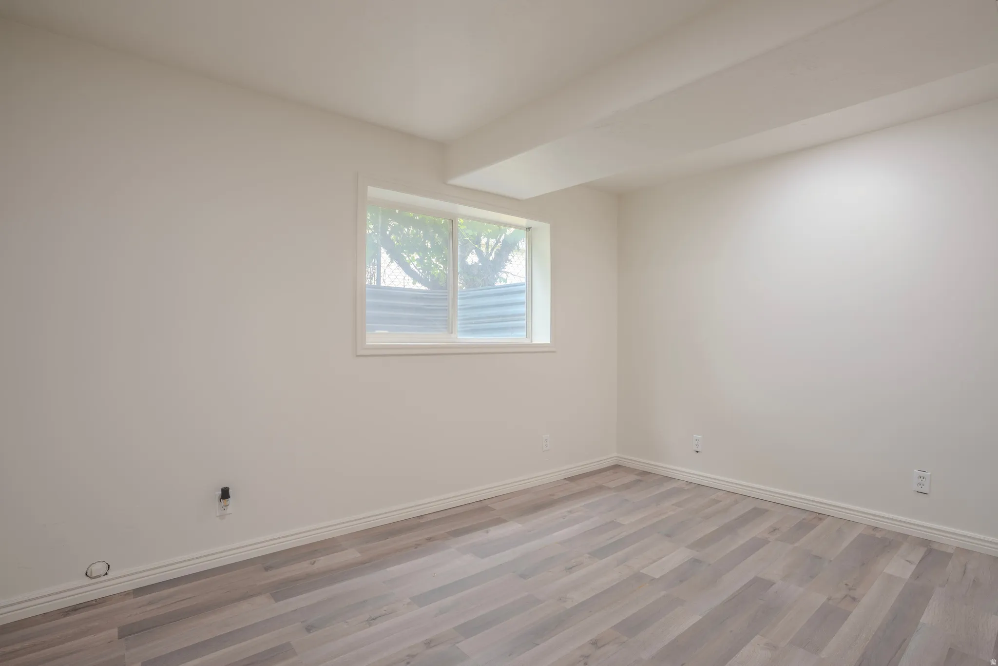 Spacious interior room featuring light wood-finish flooring, white walls, and a window with white trim