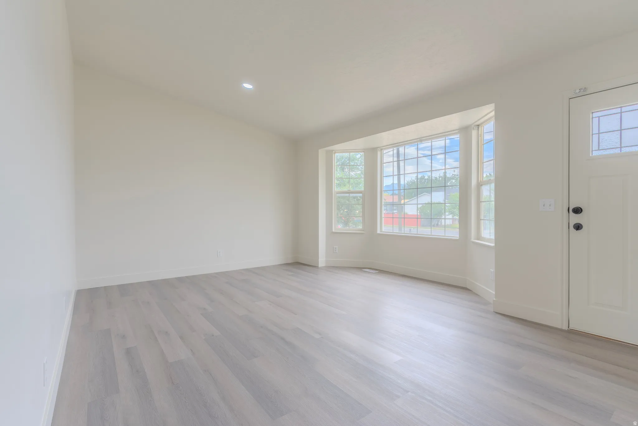 Spacious interior featuring bay windows with grid patterns, light wood-finish flooring, recessed ceiling lighting, and a paneled entry door with frosted glass