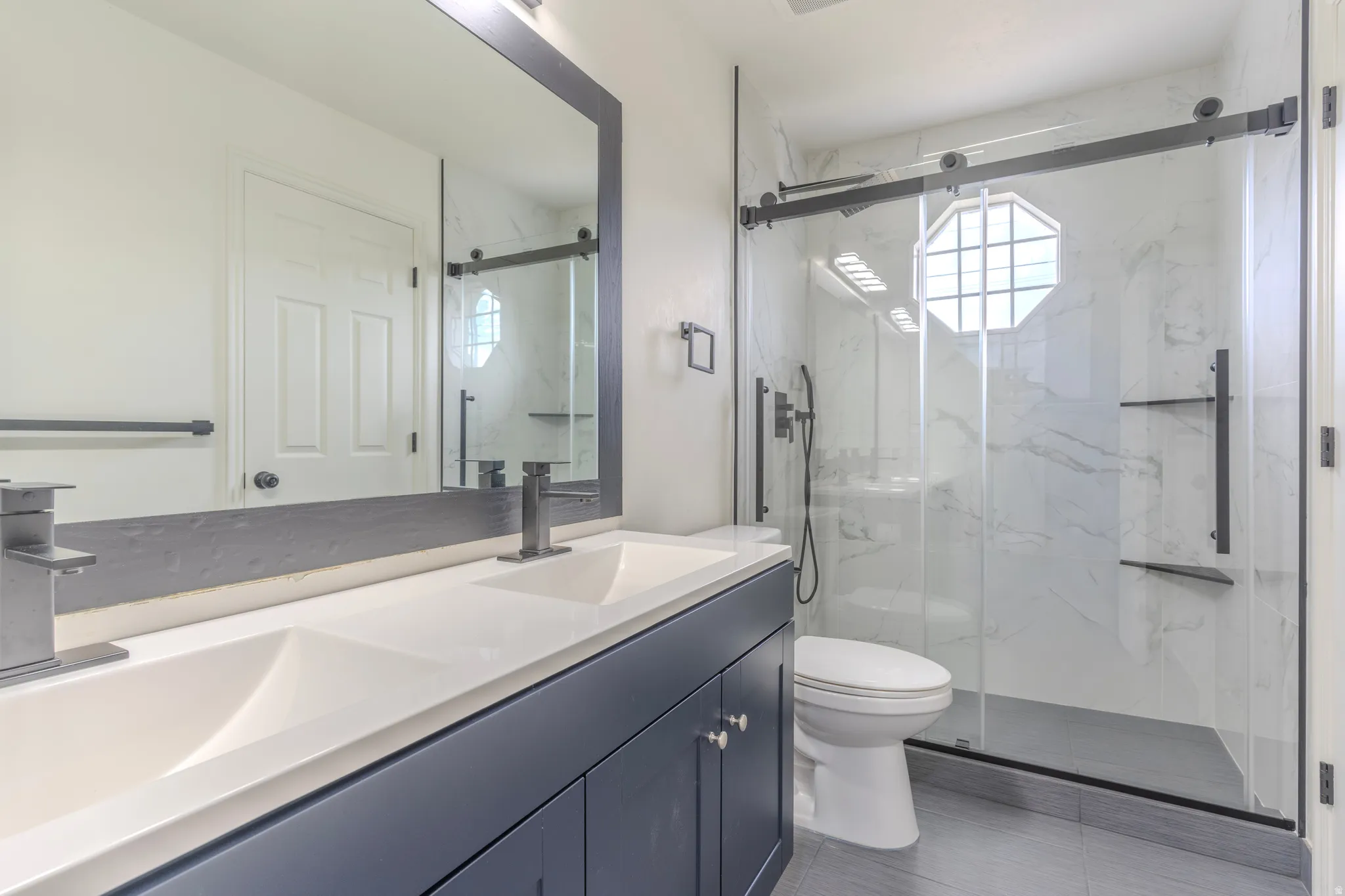 Dual vanity with white undermount sinks, flanked by a glass-enclosed shower featuring a sliding door, white marble-look tile, and a decorative octagonal window