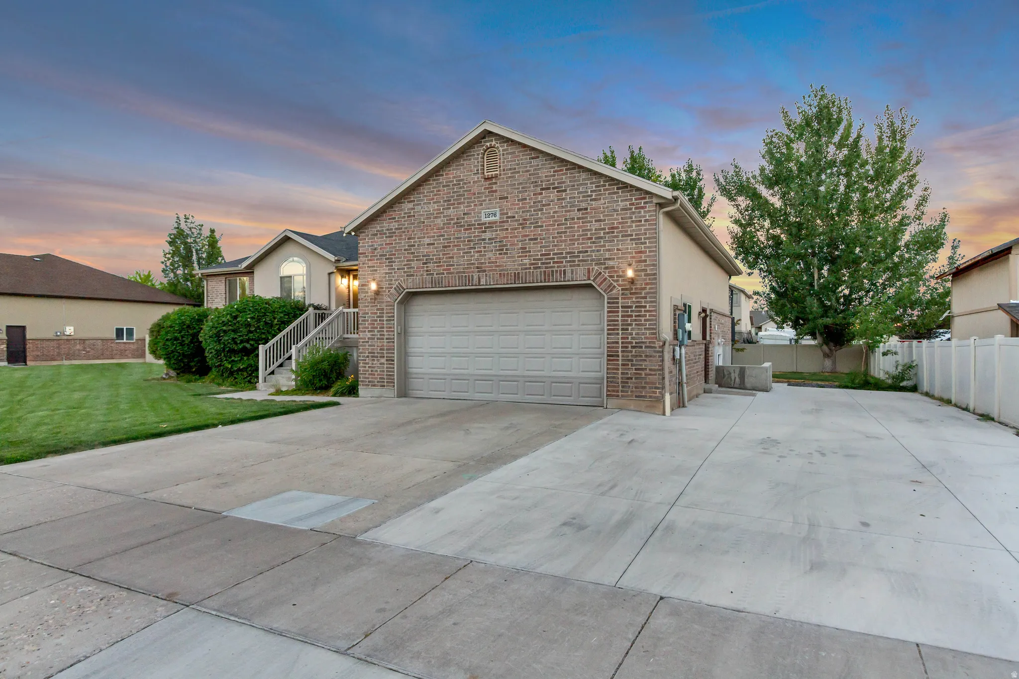 Brick facade property with an attached two-car garage, extended concrete driveway, mature landscaping, and a privacy fence
