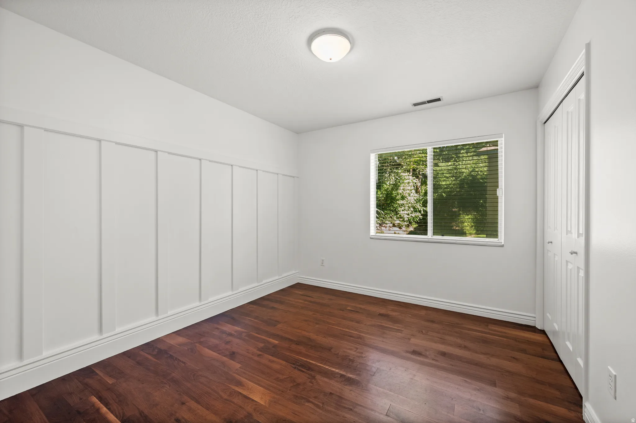 Rich wood-finish flooring, accent wall with vertical paneling, and a dual-pane window with blinds