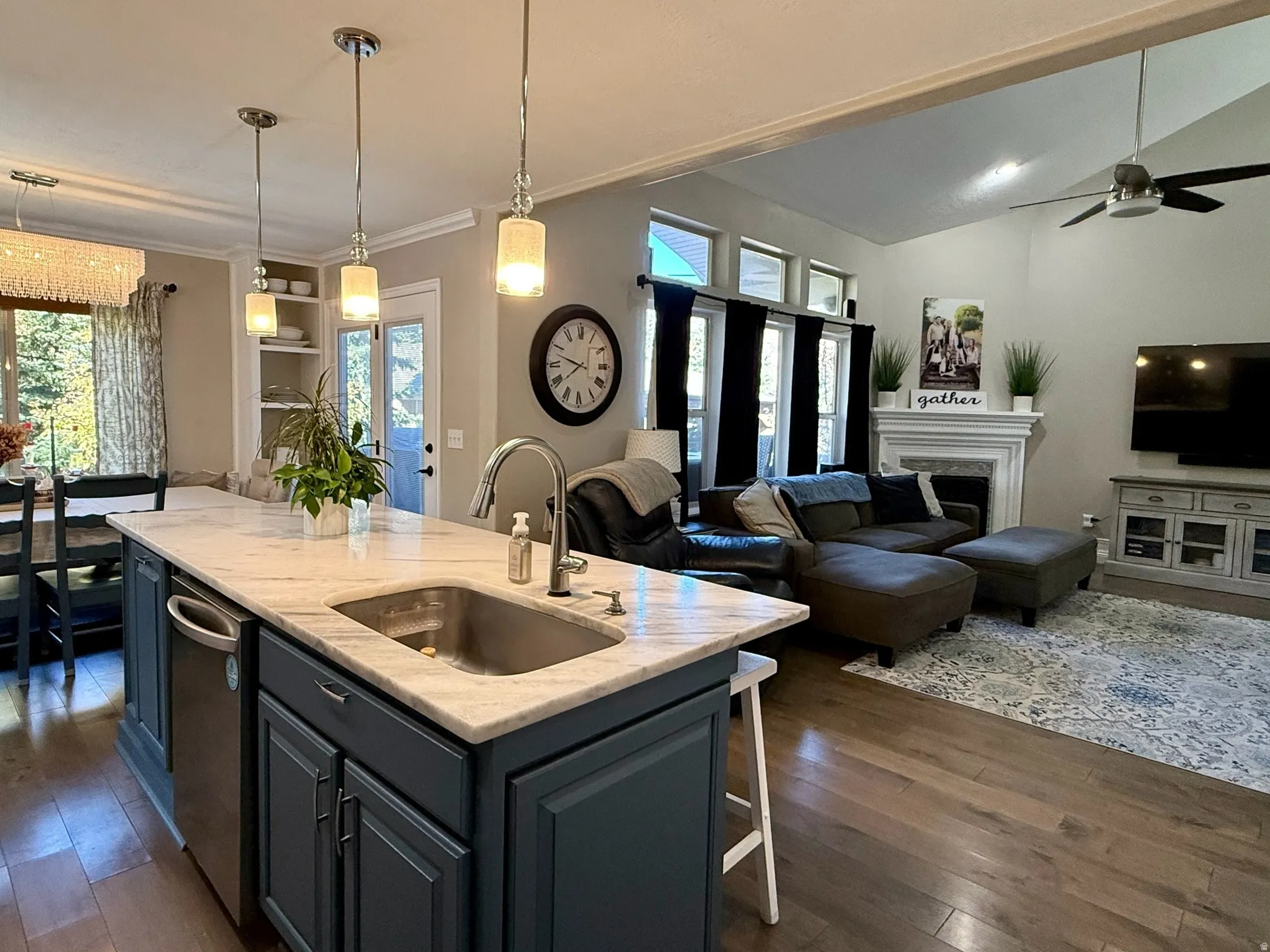 Kitchen island with a built-in sink and dark cabinetry, pendant lighting, wood-finish flooring, a vaulted ceiling, and a fireplace with a mantel