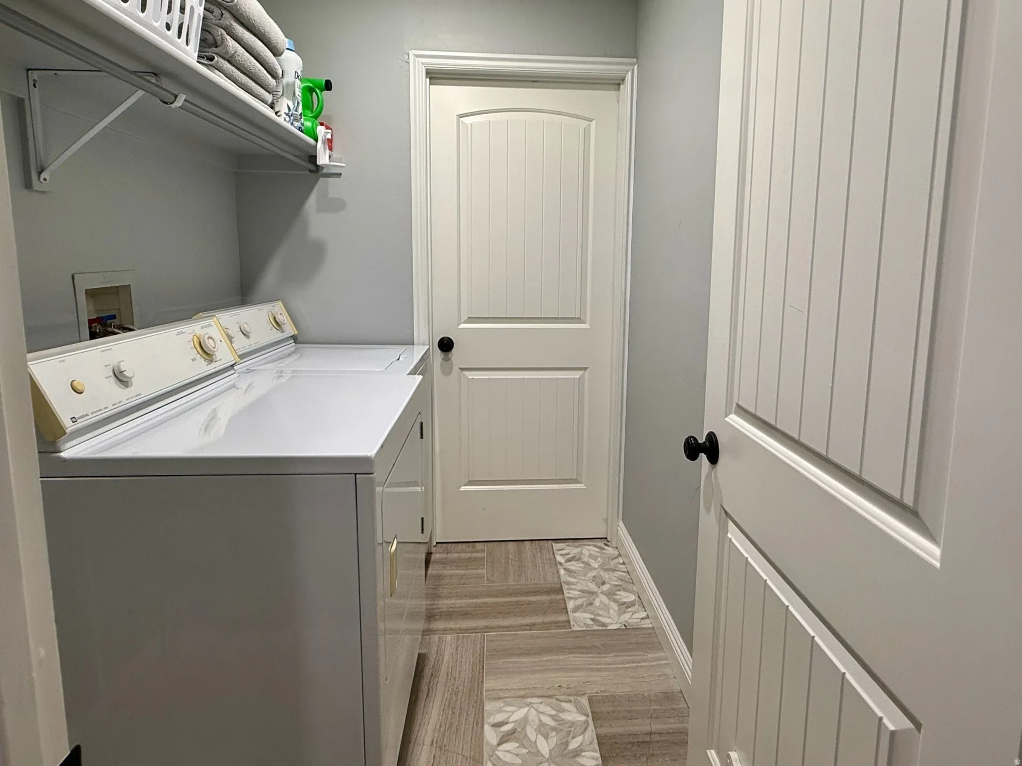 2nd Laundry in Basement-Dedicated laundry area featuring a wall-mounted shelf, light gray wall color, and wood-finish flooring with accent tiles