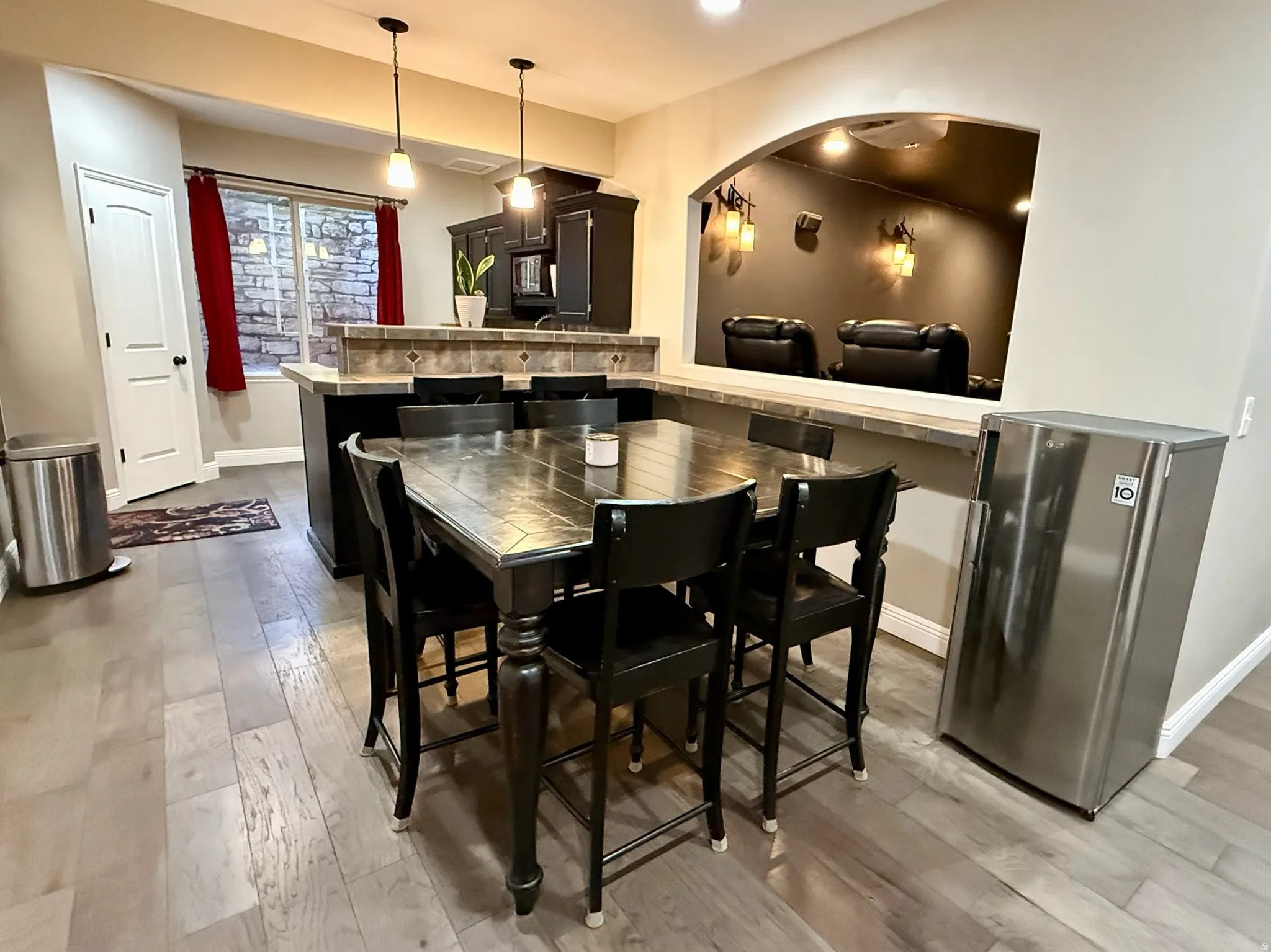 Open-concept dining area featuring wood-finish flooring, a kitchen island with a tiled backsplash and pendant lighting, and an arched pass-through to a dedicated media space
