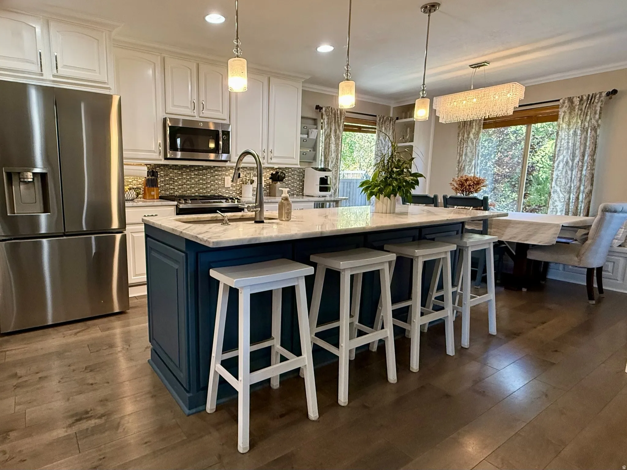 Open-concept kitchen featuring a spacious island with a marble-finish countertop, pendant lighting, wood-finish flooring, stainless steel appliances, and built-in white cabinetry