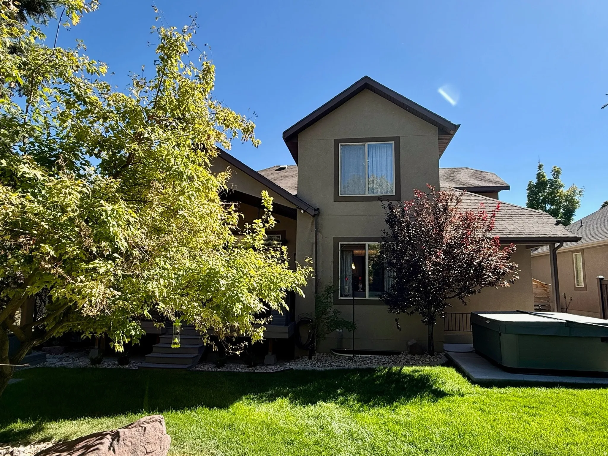 Rear exterior featuring a spa, manicured lawn, mature trees, and covered patio