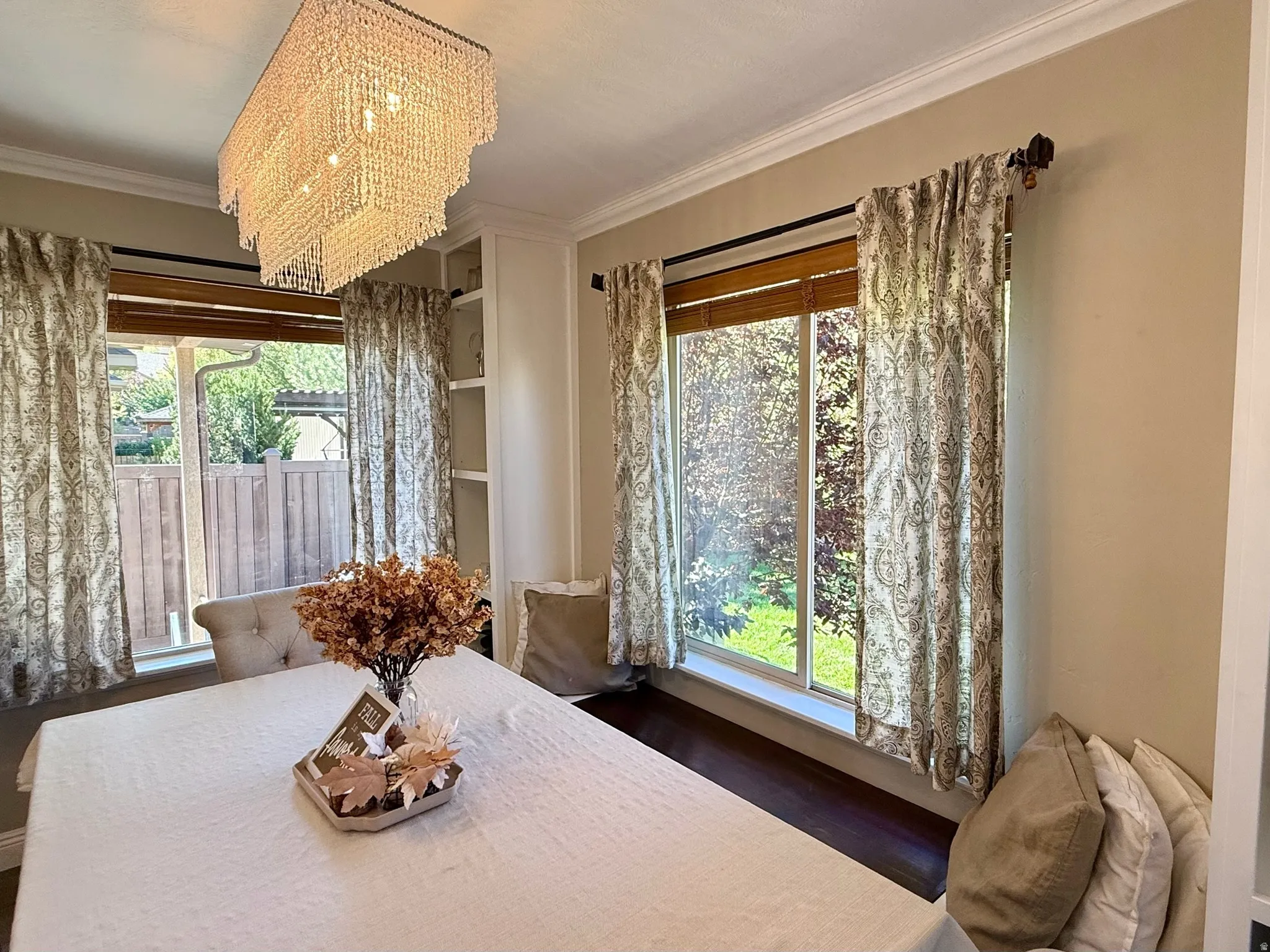 Rectangular crystal chandelier illuminating a dining area featuring built-in shelving, crown molding, and two windows with wood blinds and patterned drapes