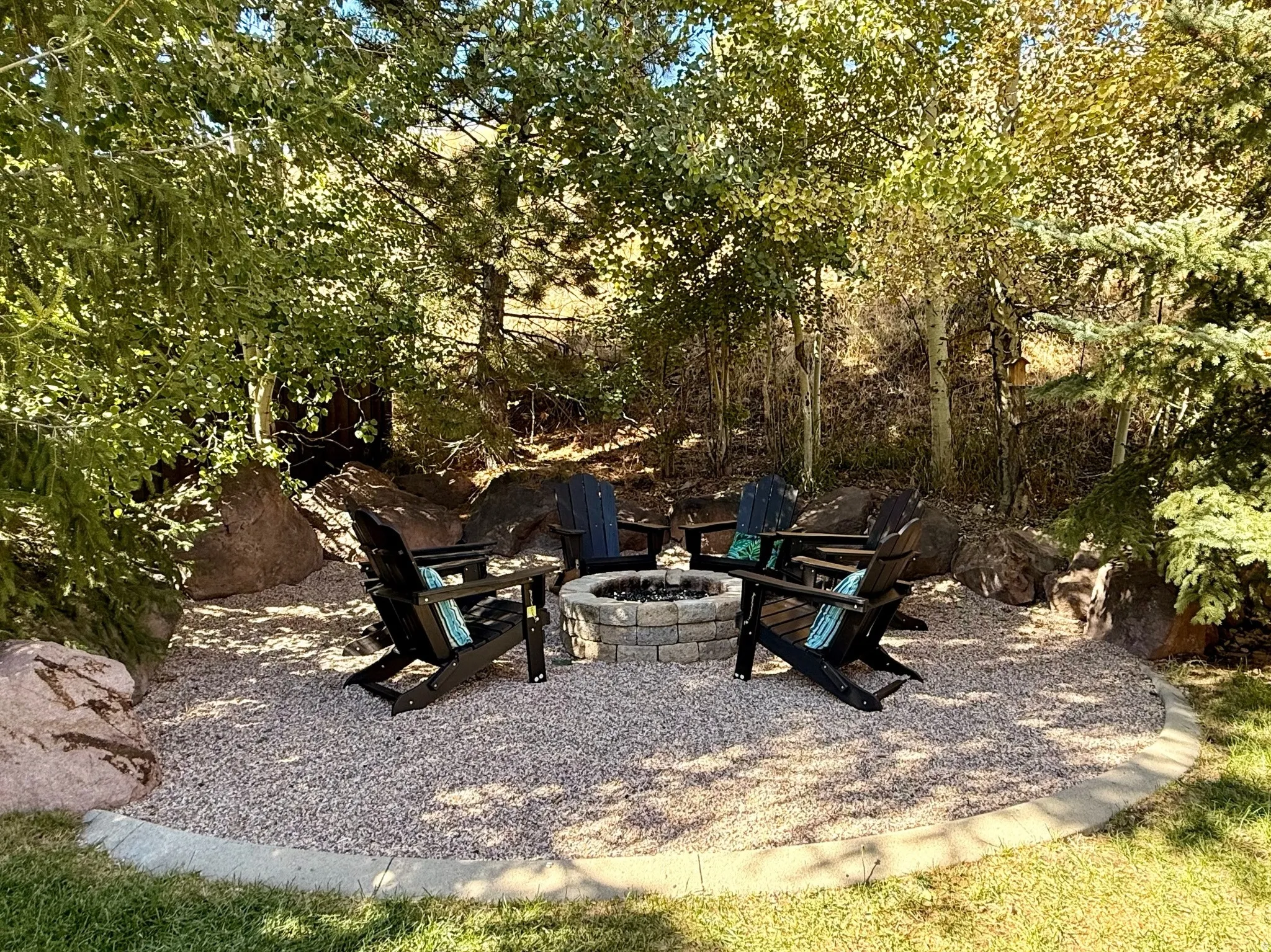 Outdoor fire pit area featuring a stone surround, gravel patio, concrete curbing, and mature trees