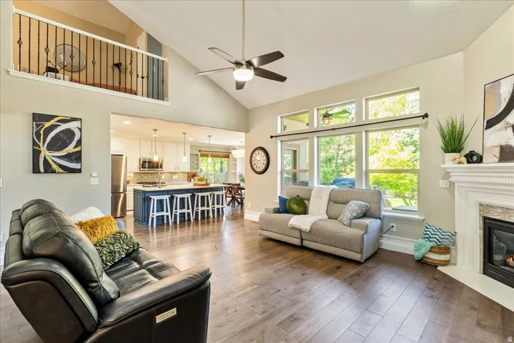 Vaulted ceiling with a ceiling fan, wood-finish flooring, fireplace with a white mantel, and a kitchen with an island and pendant lighting