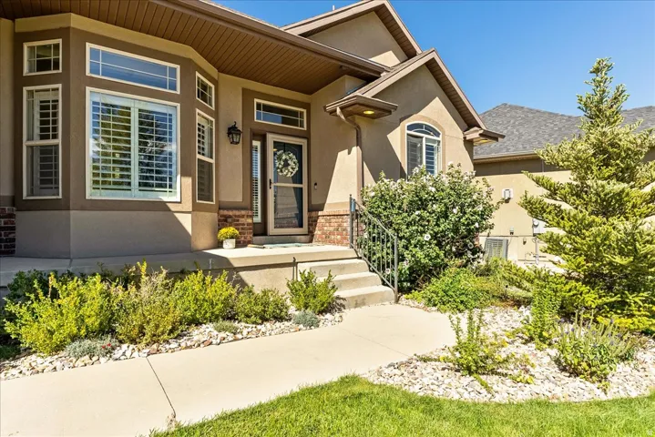 Stucco exterior with brick accents, bay window with plantation shutters, arched window with plantation shutters, concrete pathway, and landscaped garden beds
