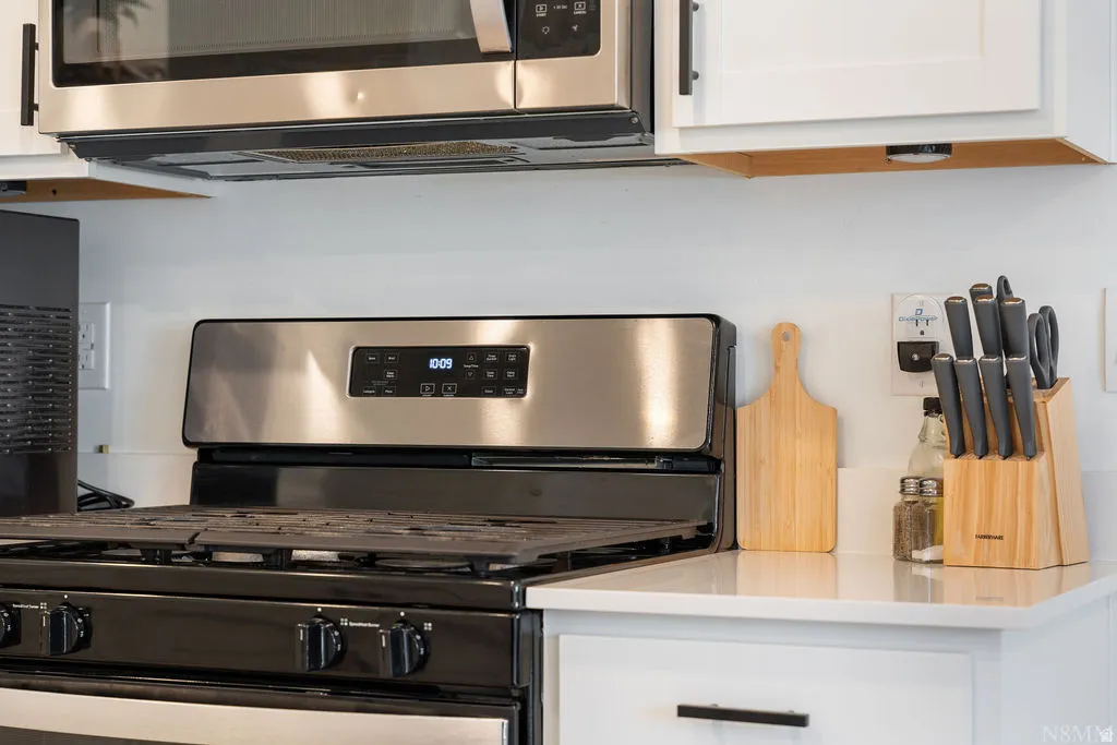 Stainless steel microwave with range hood, black gas range with grates, white cabinetry, and light-toned countertop