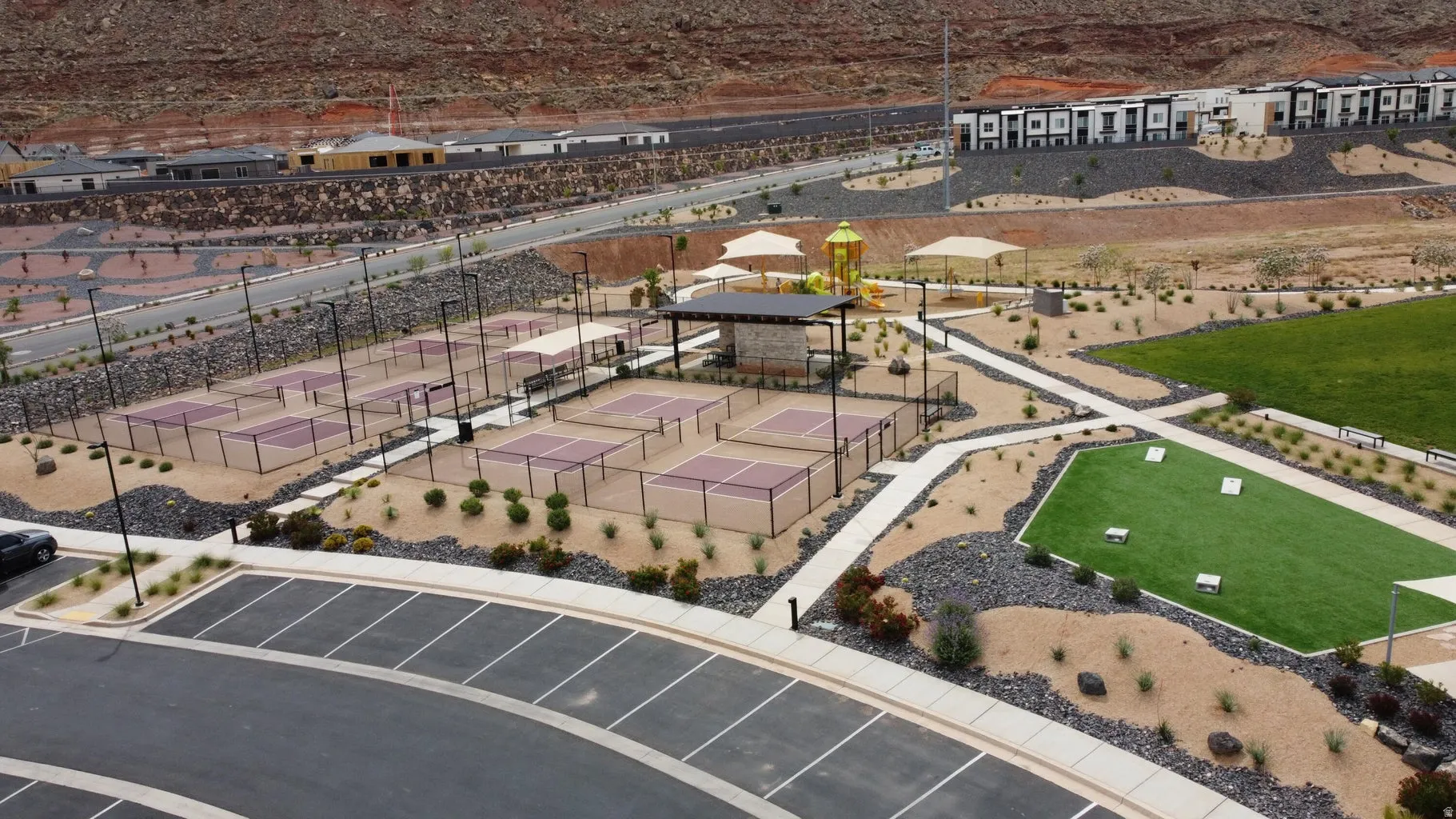 Community amenity area featuring multiple pickleball courts, a central shaded pavilion, a children's play structure with shade canopies, an artificial turf recreation area, and extensive hardscaping with desert landscaping