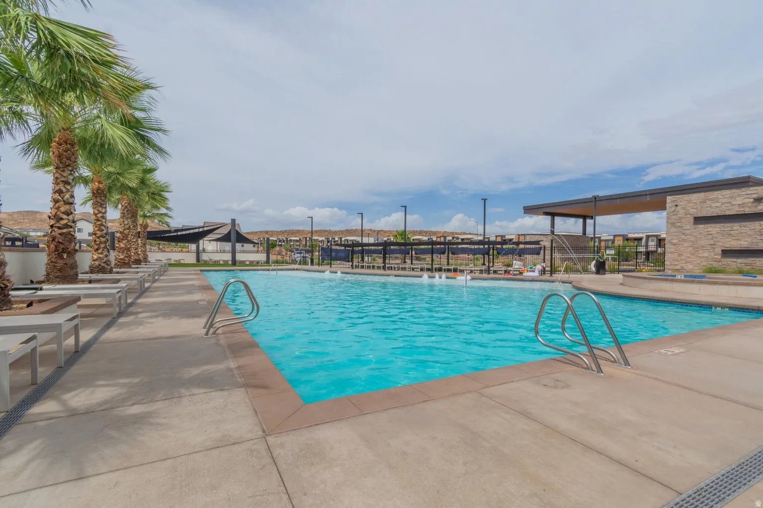 Resort-style swimming pool featuring a large concrete deck, integrated sun loungers, a stone-clad building with an extended overhang, and mature palm trees
