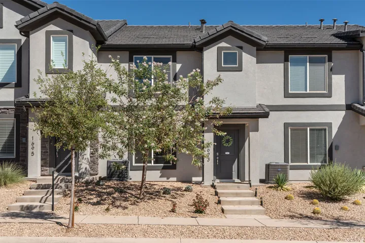 Gray stucco exterior with dark trim accents, featuring a dark gray front door, multiple windows with blinds, and mature desert landscaping