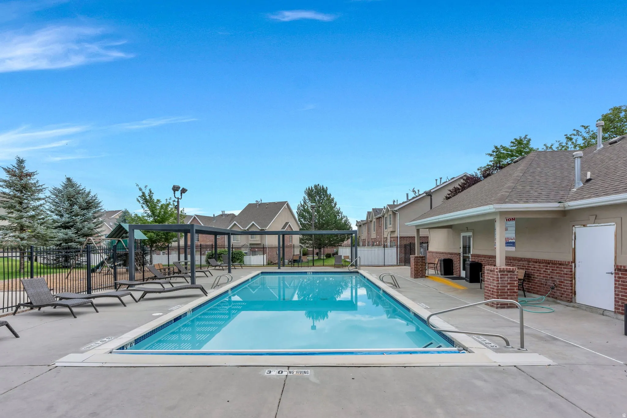 Outdoor swimming pool with concrete decking, surrounded by a metal fence and bordered by a brick and siding building