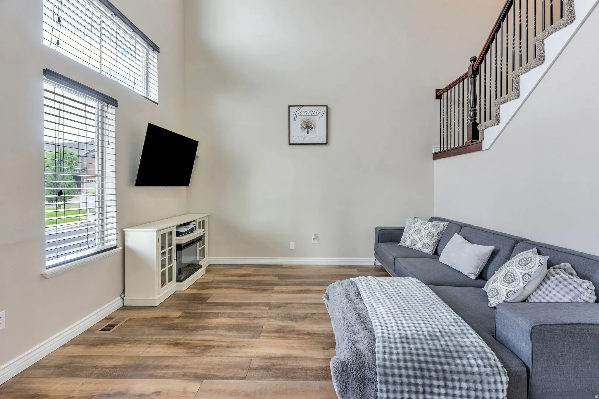 Living area with wood-finish flooring, high ceilings, large windows with blinds, a built-in electric fireplace media console, and a staircase with wood newel posts and banister