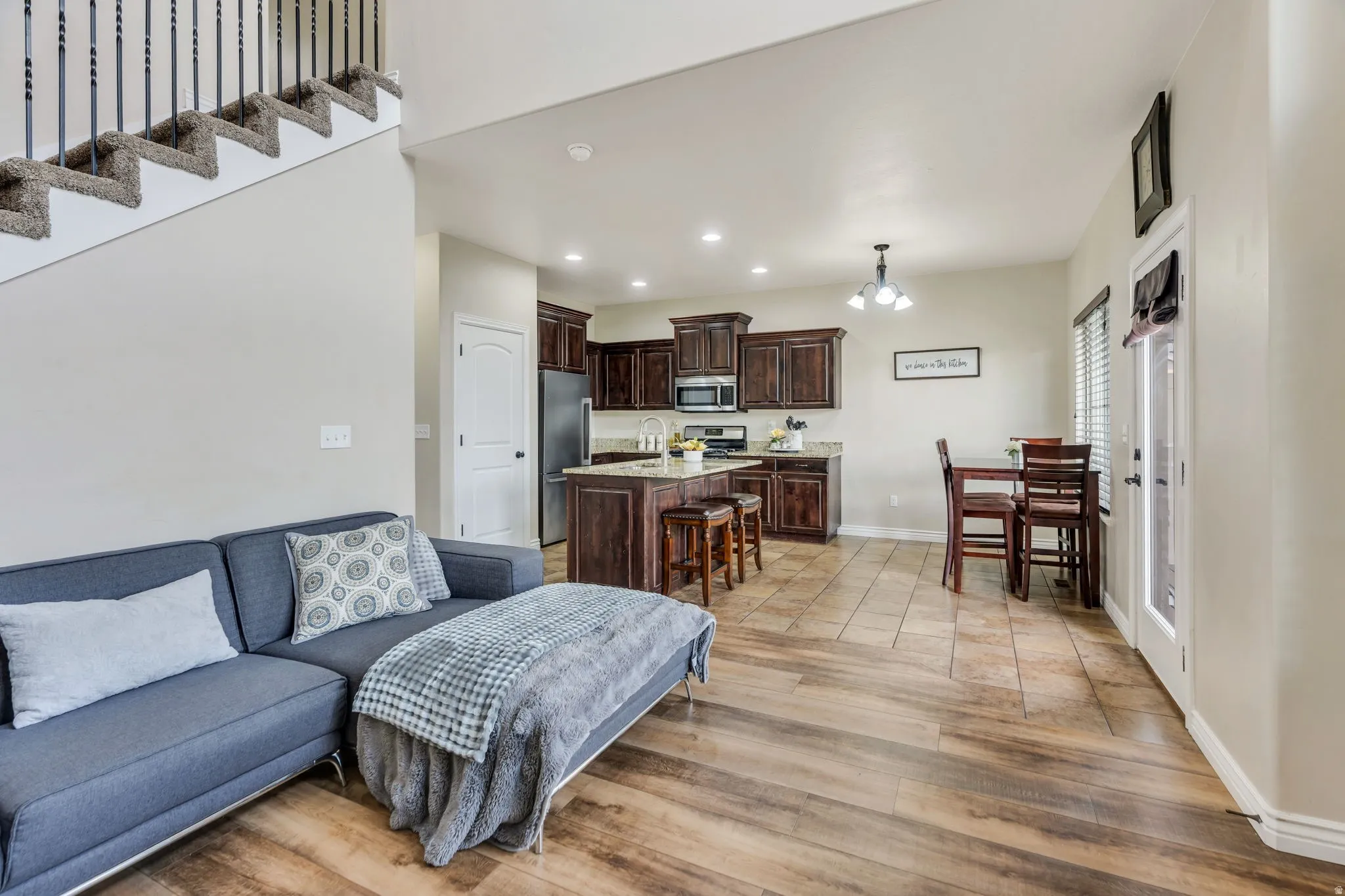 Open-concept living area featuring wood-finish flooring, a carpeted staircase with dark iron balusters, and a kitchen with dark wood cabinetry