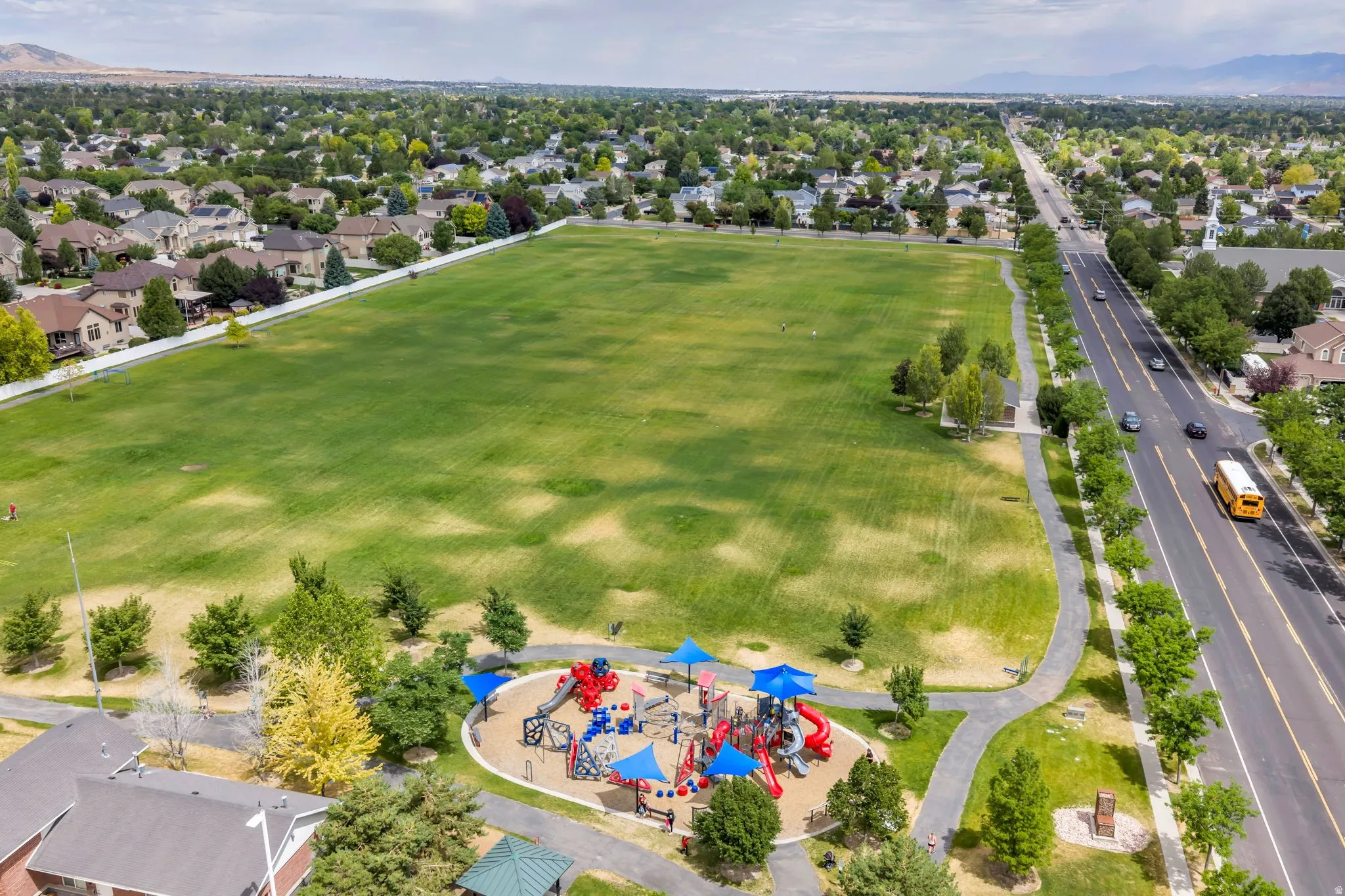 Expansive green park space featuring a modern playground with multiple slides and climbing structures, shaded by blue canopies