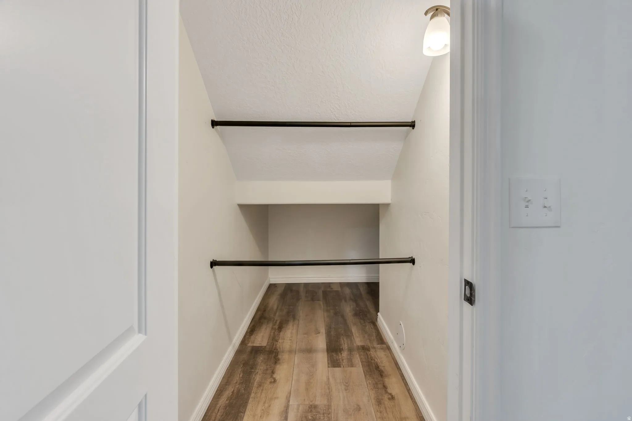 Angled storage closet featuring wood-finish flooring, two black metal hanging rods, a built-in shelf, and ceiling-mounted lighting