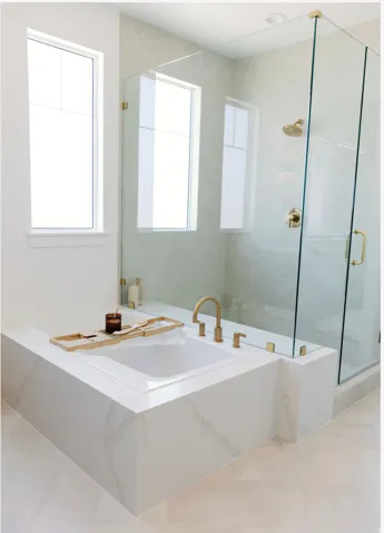 Integrated soaking tub with a white stone surround, brass fixtures, and an adjacent glass-enclosed shower with matching brass hardware and stone-look wall tile