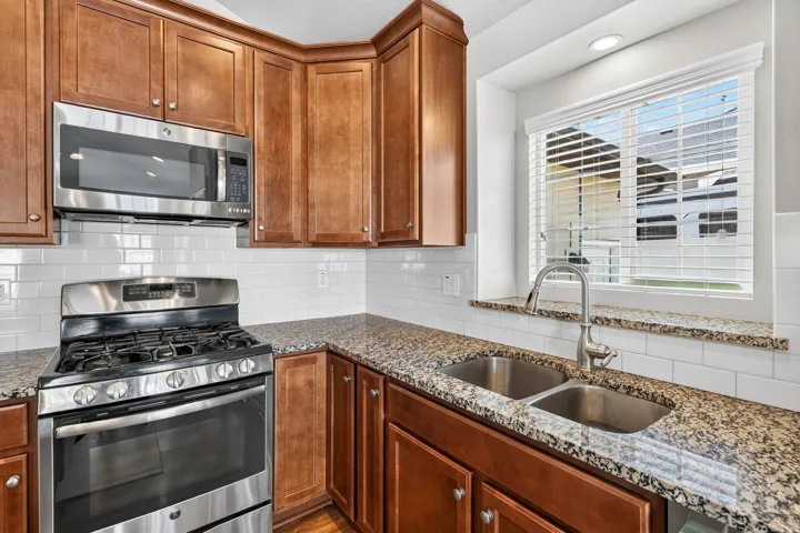 Kitchen featuring stainless steel appliances, wood cabinetry, granite countertops, a double basin sink with a gooseneck faucet, and white subway tile backsplash