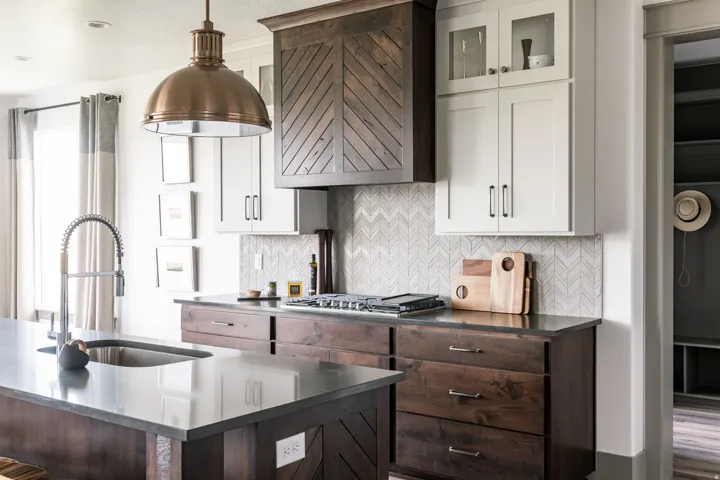 Kitchen featuring a brass pendant light, a dark wood range hood with a chevron pattern, light gray upper cabinetry, dark wood lower cabinetry, and a light-toned herringbone backsplash