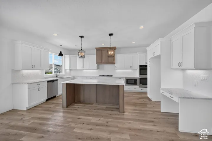 Spacious kitchen featuring an expansive island with a stone-finish countertop, white shaker-style cabinetry, stainless steel appliances, wood-finish flooring, and a decorative range hood