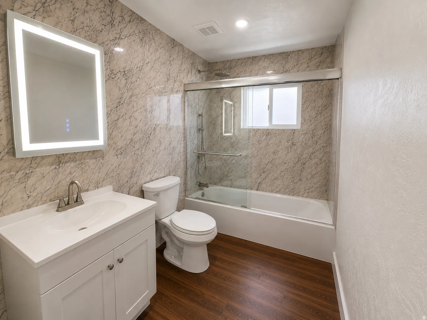 Modern bathroom featuring an illuminated mirror, white vanity with integrated sink, wood-finish flooring, a toilet, and a bathtub with a sliding glass enclosure