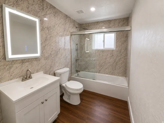 Modern bathroom featuring an illuminated mirror, white vanity with integrated sink, wood-finish flooring, a toilet, and a bathtub with a sliding glass enclosure