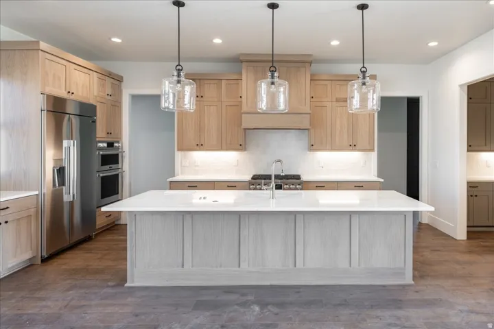 Kitchen featuring a large central island with a sink, stainless steel appliances including a refrigerator and double ovens, light wood cabinetry, a tile backsplash, and three pendant lights