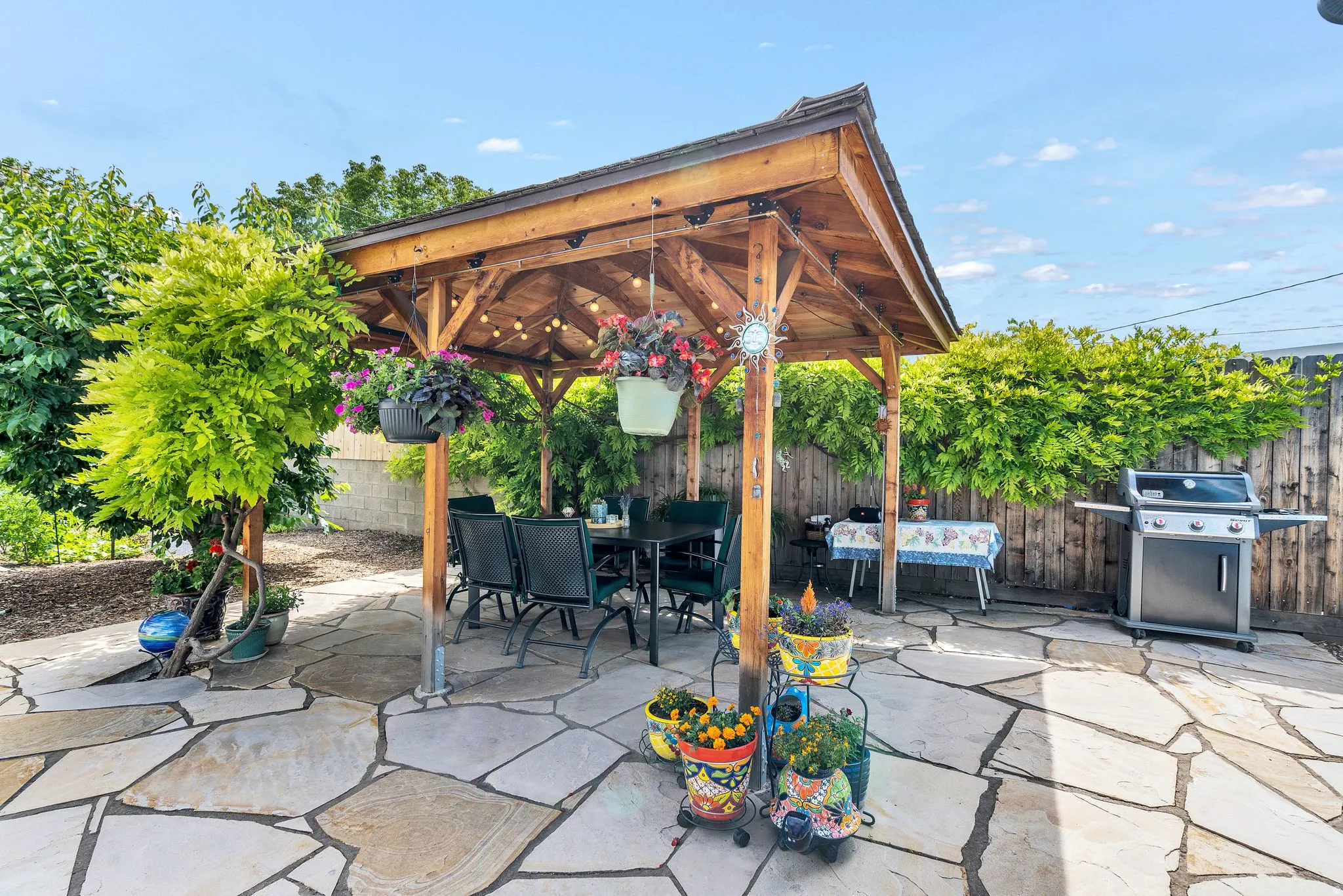 Spacious flagstone patio featuring a wooden pergola with string lighting, surrounded by mature trees and a privacy fence