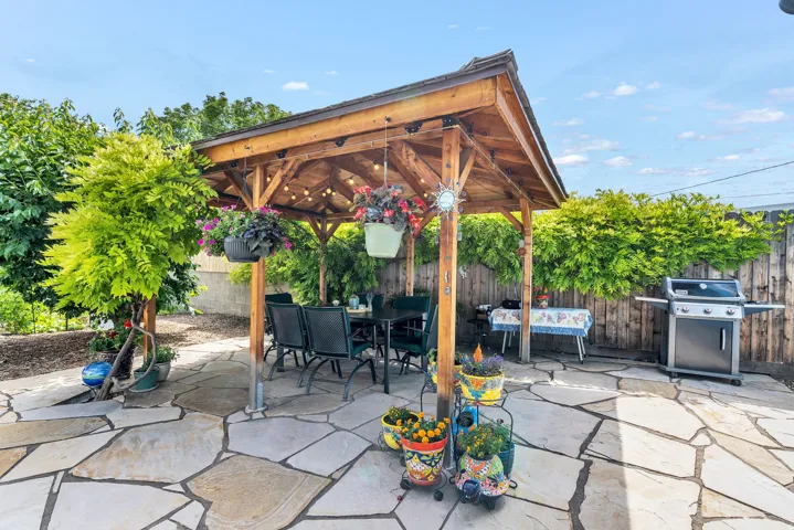 Spacious flagstone patio featuring a wooden pergola with string lighting, surrounded by mature trees and a privacy fence
