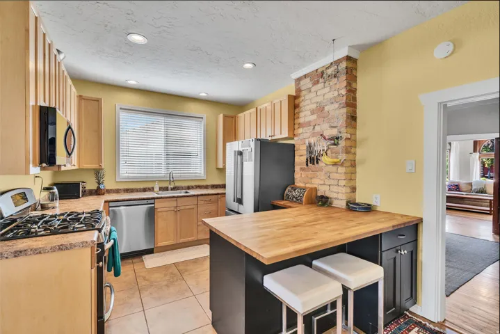 Kitchen featuring a original brick accent wall, light wood cabinetry, stainless steel appliances, a center island with a wood countertops, and tile flooring