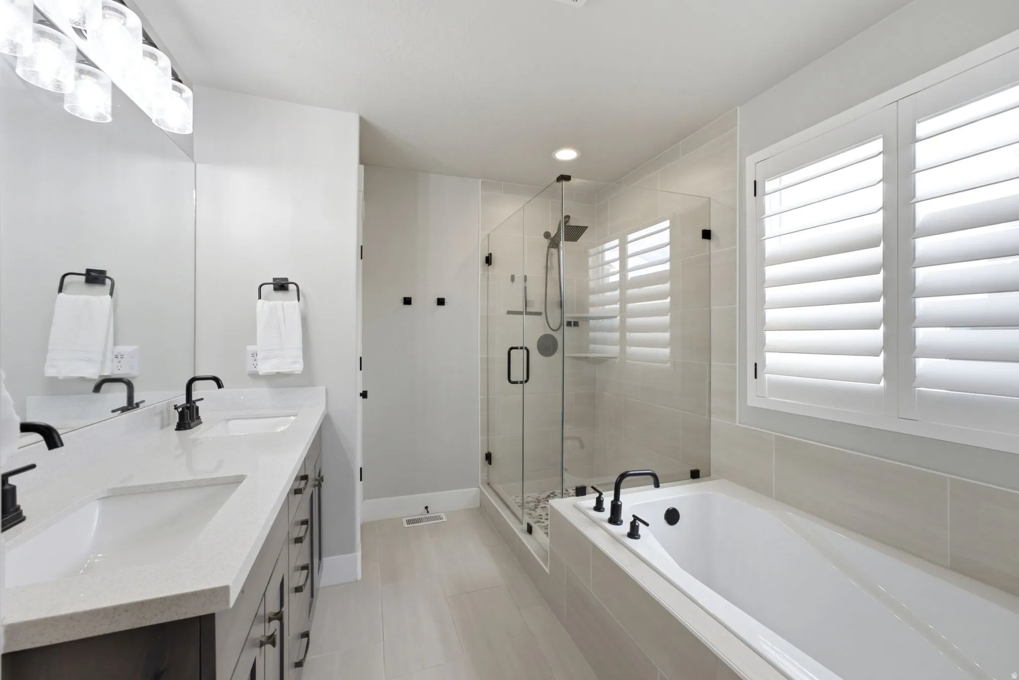 Contemporary bathroom featuring a spacious walk-in shower with a glass enclosure, a white soaking tub with matte black fixtures, and dual sinks set in a light-colored countertop