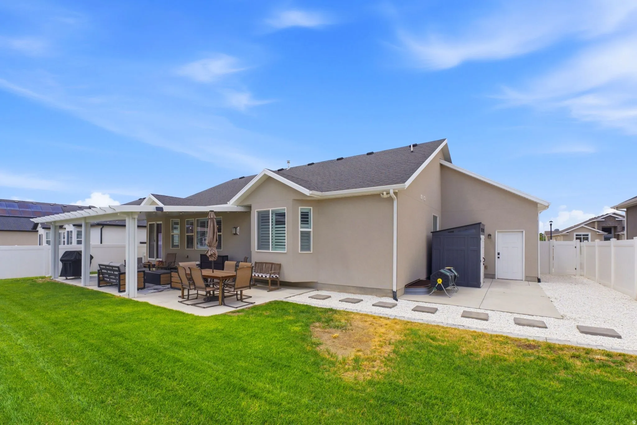 Expansive backyard featuring a concrete patio with an attached white pergola, a manicured lawn, white privacy fencing, and a gravel side yard with stepping stones