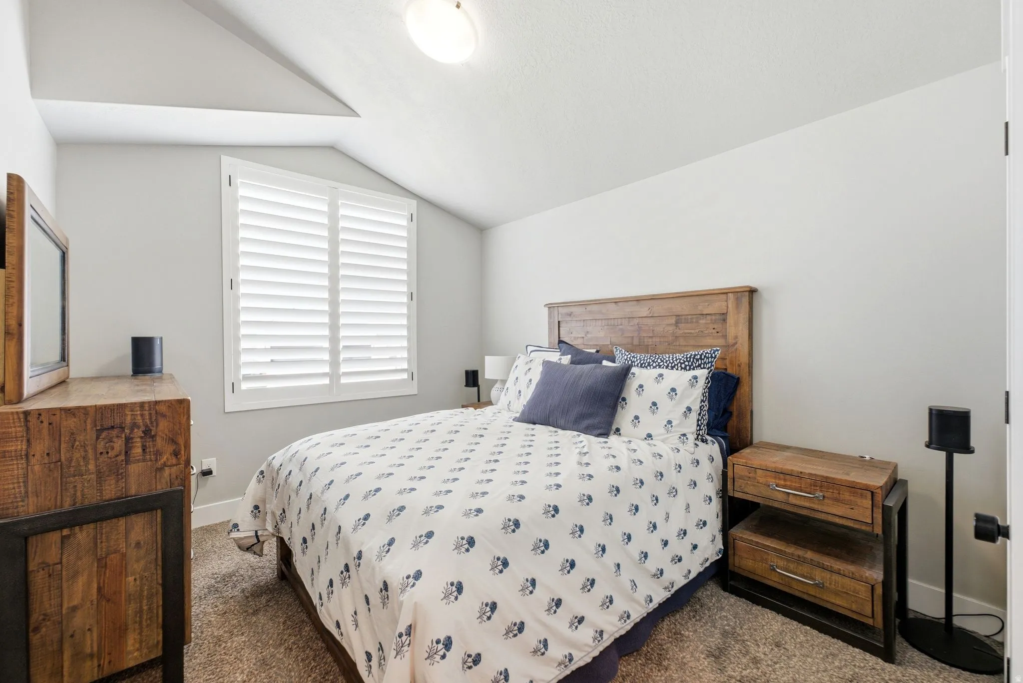 Bedroom featuring a sloped ceiling, recessed window with plantation shutters, wood-finish headboard, and wood-finish bedside table with metal accents