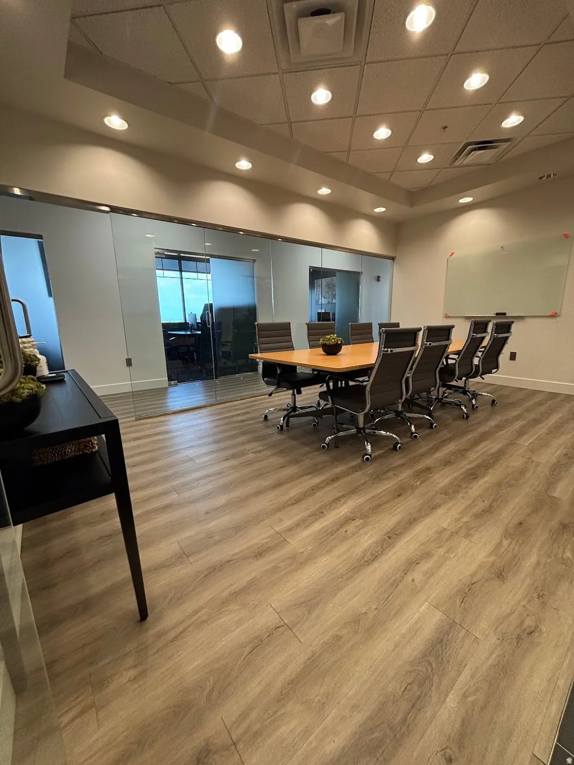 Conference room featuring glass panel walls, wood-finish flooring, a recessed ceiling with integrated lighting, and a wall-mounted whiteboard
