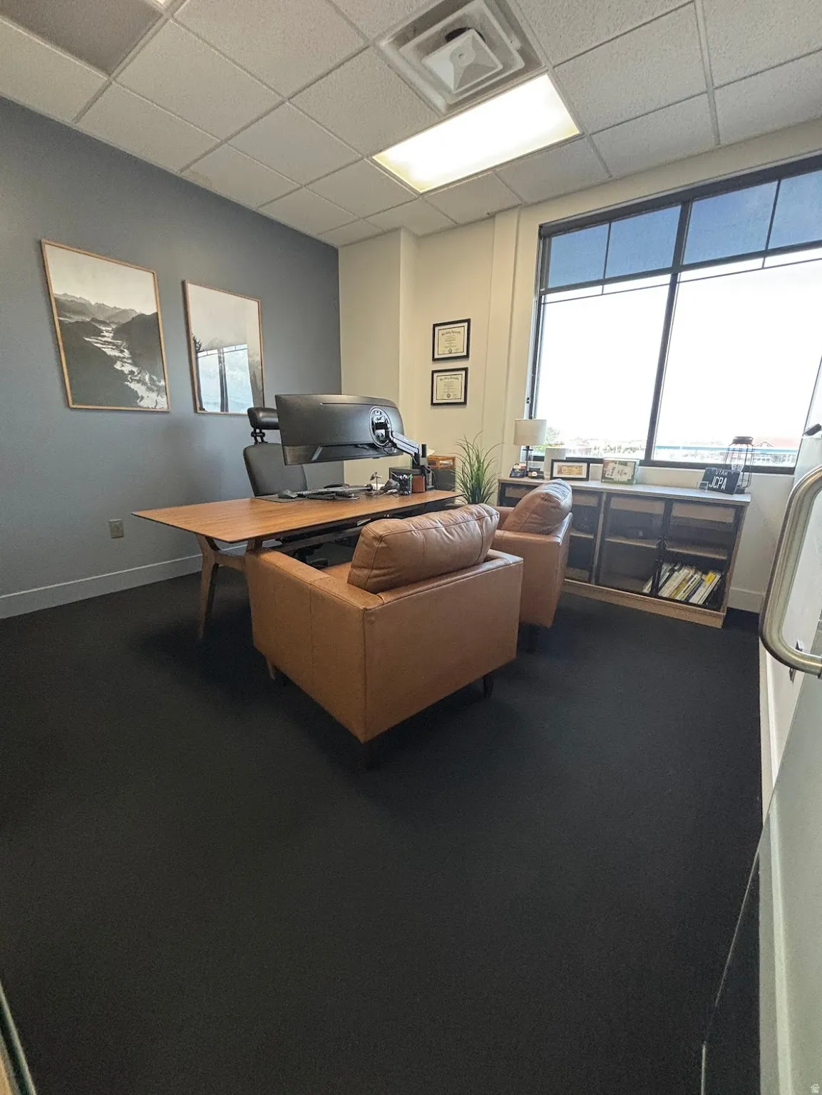 Private office space featuring a large window with roller shades, dark wall accent, recessed lighting, and dark carpeting.