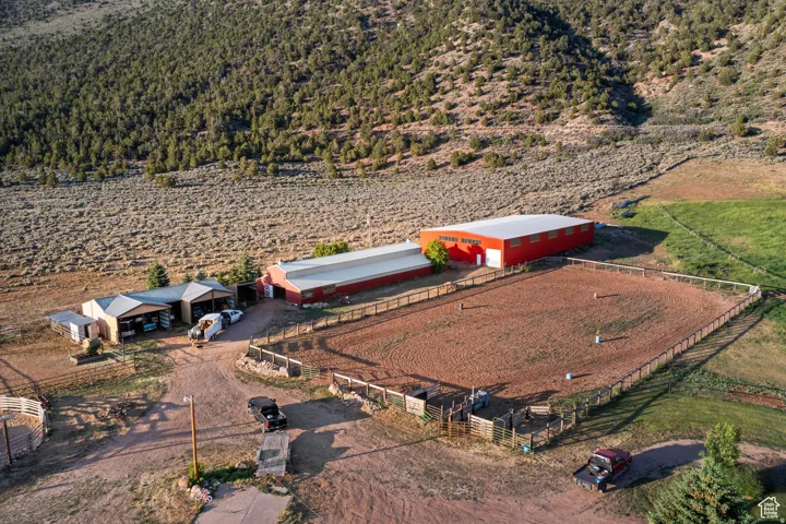 Expansive rural property featuring multiple outbuildings, a large red barn with a metal roof, an outdoor arena with fencing, and a treed hillside backdrop